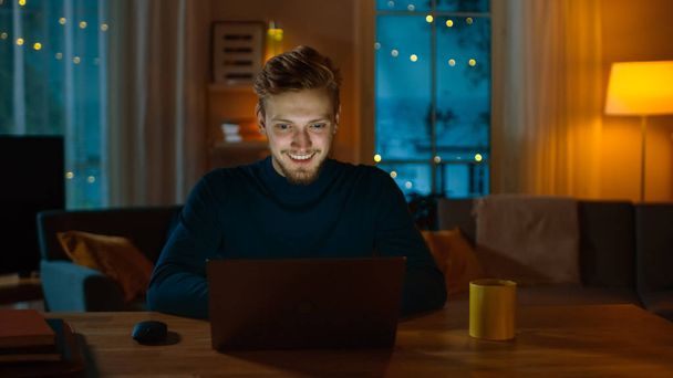 Handsome Smiling Man Works on a Laptop while Sitting at His Desk at Home. Young Freelancer Works on Computer in His Cozy Living Room with Warm Evening Lighting Turned on. - Photo, Image