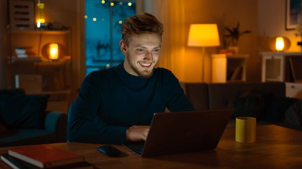 Handsome Smiling Man Works on a Laptop Computer while Sitting at His Desk at Home. Cozy Living Room with Warm Evening Lighting Turned on. - Photo, Image