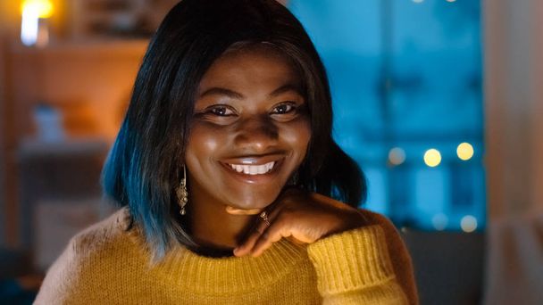Portrait of Beautiful Black Girl Uses Laptop while Sitting at Her Desk at Home, Her Face is Illuminated with a Screen Light. In the Cute Young Woman Smiles on Camera. - Photo, Image