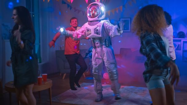 カレッジハウスの衣装パーティーで:宇宙服を着た楽しい男はオフに踊り、ロボットダンスモダンな動きを行います。彼と一緒にネオンライトで踊る美しい女の子と男の子. - 写真・画像