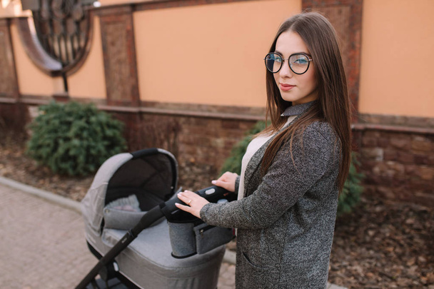 Young mother walking with her newborn baby in stroller. Grey pram. Beautiful mom in sunglasses - 写真・画像