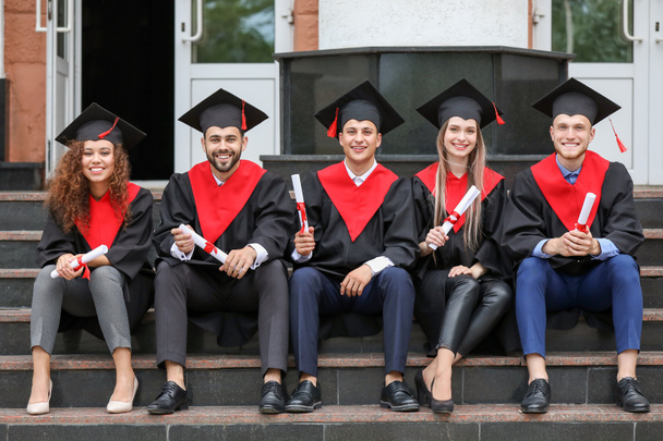Jonge studenten in Bachelor gewaden en met diploma's zittend op de trap buitenshuis - Foto, afbeelding