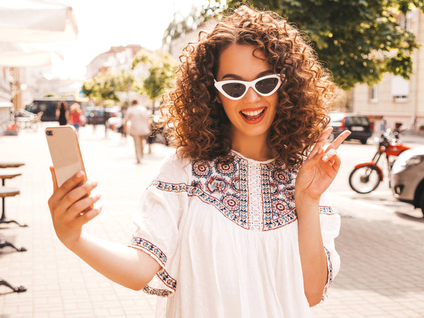 Beautiful smiling model with afro curls hairstyle dressed in summer hipster white dress.Sexy carefree girl posing in the street in sunglasses.Taking selfie self portrait photos on smartphone - Foto, Bild