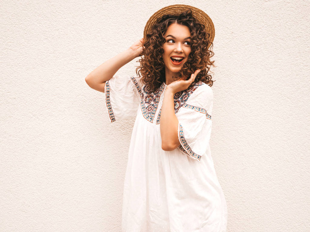 Piękny uśmiechnięty model z Afro loki fryzura ubrana w letni Hipster Biała Sukienka. Seksowna beztroza dziewczyna pozowanie na ulicy w pobliżu białej ścianie w kapeluszu. Zabawna, pozytywna kobieta zabawy i szaleje - Zdjęcie, obraz