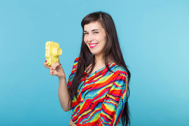 Smiling young woman is holding in her hands a yellow vintage camera model posing on a blue background. Concept of amateur and professional photography. - Foto, imagen