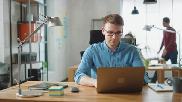 Handsome Young Man in Glasses and Shirt is Working on a Laptop in a Creative Business Agency. They Work in Loft Office. Diverse People Working in the Background. He's in Good Mood. - Záběry, video
