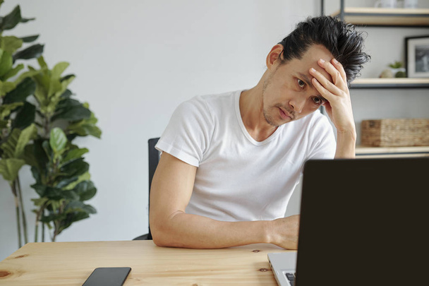 Muotokuva tylsistynyt tai stressaantunut aasialainen mies työskentelee kannettavan toimiston pöydässä
 - Valokuva, kuva