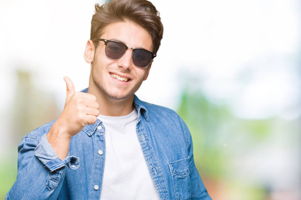 Jonge knappe man dragen van een zonnebril op geïsoleerde achtergrond gelukkig duimschroef opwaarts gebaar met de hand te doen. Goedkeuring van de expressie te kijken naar de camera toont succes. - Foto, afbeelding