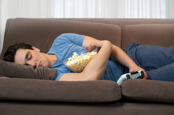 junger gutaussehender Mann schläft ein, nachdem er faszinierendes Videospiel gespielt hat, bei dem er den Steuerknüppel in der einen und Popcorn in der anderen Hand hält - Foto, Bild