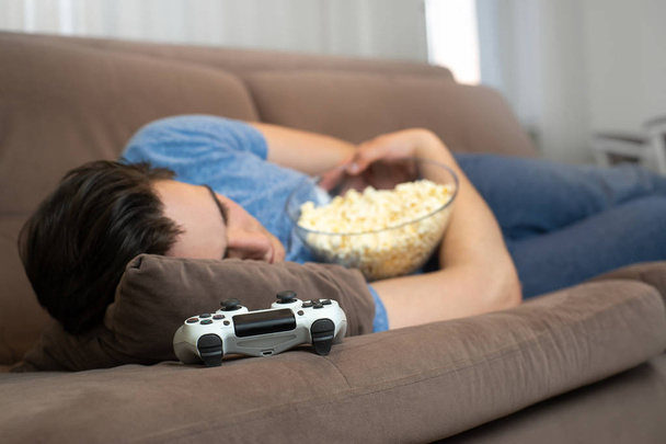 young handsome man fall asleep after playing fascinating video game still holding popcorn in his hands - Foto, Bild