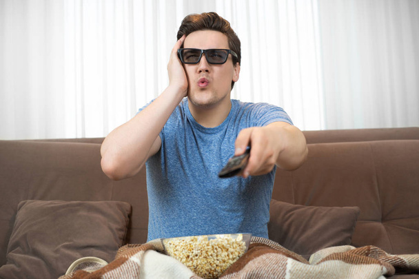 young surprised man wearing sunglasses watching movie eating popcorn clicking remote control looks astonished - Фото, изображение