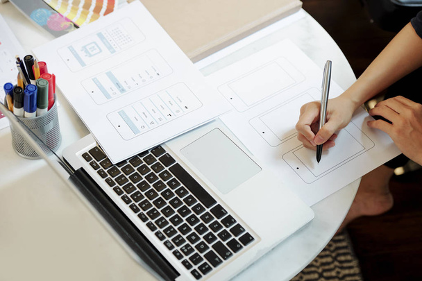 Крупный план женщины-дизайнера, сидящей за столом с ноутбуком и рисующей эскиз мобильного телефона на бумаге
 - Фото, изображение
