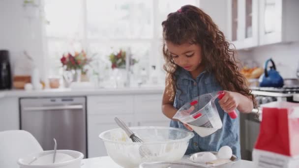 Young girl in the kitchen preparing cake mixture on her own, adding milk and mixing, front view - Felvétel, videó