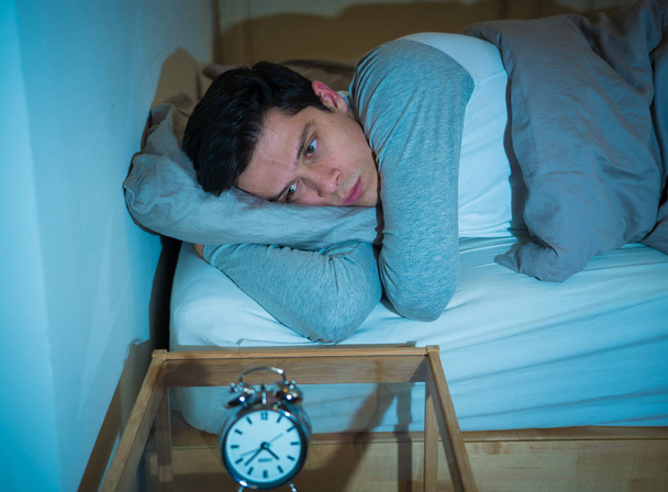 Sleepless and desperate young caucasian man awake at night not able to sleep, feeling frustrated and worried looking at clock suffering from insomnia in stress and sleep disorder concept. - Photo, Image