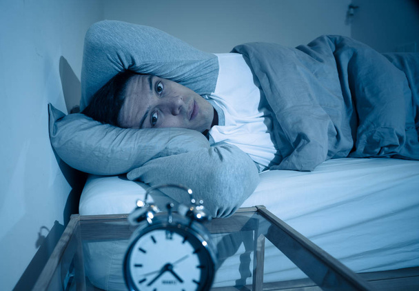 Άγρυπνος και απελπισμένος νέος Καυκάσιος άνθρωπος ξύπνιος τη νύχτα δεν είναι σε θέση να κοιμηθεί, νιώθοντας απογοητευμένος και ανήσυχος κοιτάζοντας το ρολόι που πάσχουν από αϋπνία σε άγχος και διαταραχή ύπνου αντίληψη. - Φωτογραφία, εικόνα