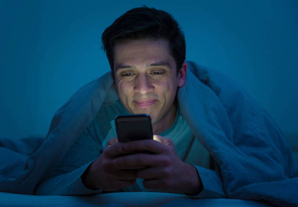Young mobile phone addict man awake at night in bed using smartphone for chatting, flirting and sending text message. In communication, Internet addiction and Social media network abuse concept - Foto, Imagen