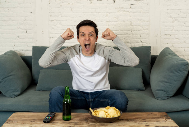 Lifestyle portrait of excited football fan having fun watching soccer or football game on television. Enjoying and celebrating goal and victory drinking beer and eating chips. Sports fans and Goal. - Zdjęcie, obraz