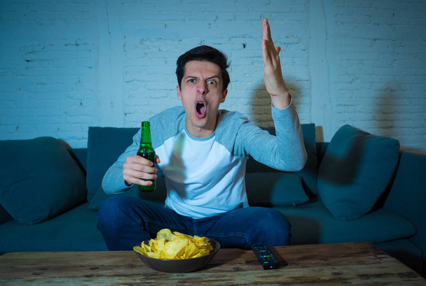 Young man drinking beer at home watching soccer football match or sports game in television at night. Screaming and celebrating goal or victory. In celebration, sports and fans concept. - Photo, image
