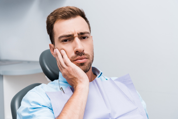 chateado barbudo homem tocando rosto enquanto tendo dor de dente na clínica dentária
  - Foto, Imagem