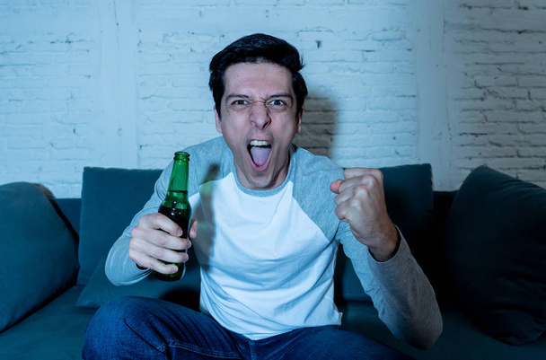 Lifestyle portrait of football fan young man with beer watching soccer football game on TV late at night. Screaming and celebrating goal or victory. In sports, people, entertainment and celebration. - Фото, изображение