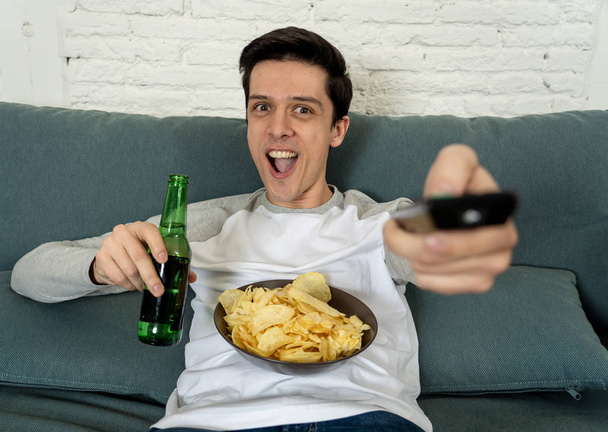Lifestyle portrait of young man on sofa watching sports or exciting movie on television. Having fun at home enjoying and celebrating goal and victory drinking beer. In entertainment and mass media. - Fotoğraf, Görsel
