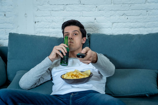 Lifestyle portrait of young bored man on couch with remote control zapping for movie or live sport. Looking disinterested drinking beer. Sedentary and mass social media or Television addiction. - Фото, изображение