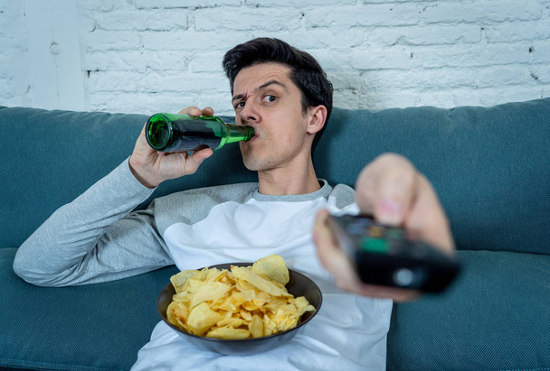 Young man switching channels in dislike feeling disgust with TV shows or movies. With a look of disapproval while drinking beer. Rubbish in and bad television shows and unhealthy lifestyle. - Fotografie, Obrázek