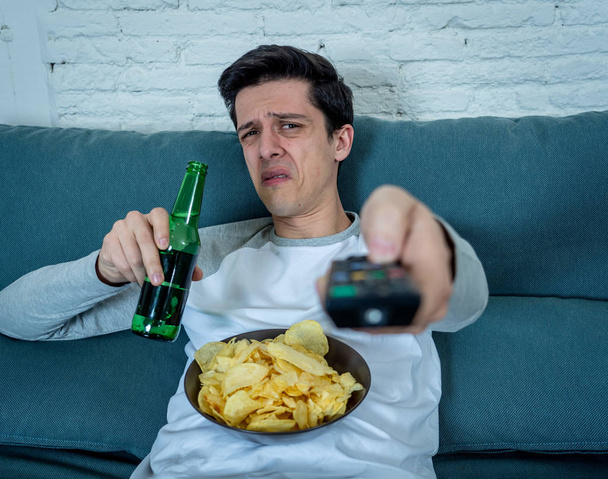 Young man switching channels in dislike feeling disgust with TV shows or movies. With a look of disapproval while drinking beer. Rubbish in and bad television shows and unhealthy lifestyle. - Fotoğraf, Görsel