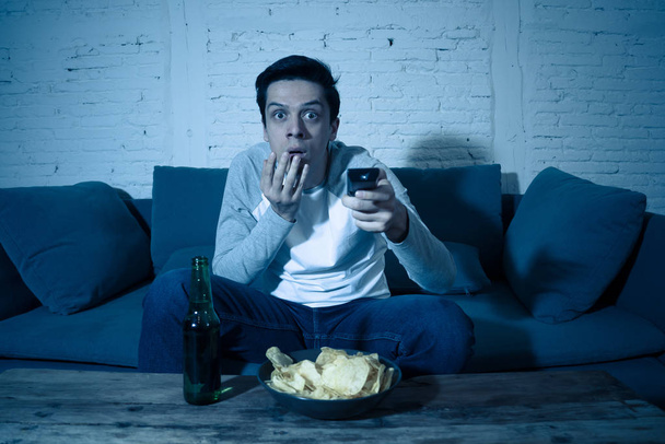 Lifestyle portrait of young scared man watching horror movie or thriller on TV. Holding remote control shocked making frightened gestures. In people, mass media, television and entertainment concept. - Photo, Image