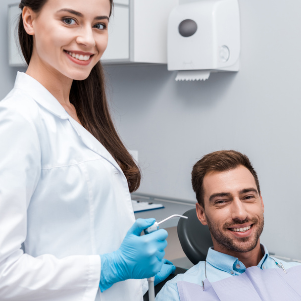 piękny i szczęśliwy dentysta posiadający sprzęt dentystyczny w pobliżu szczęśliwego mężczyzny  - Zdjęcie, obraz