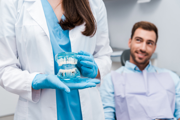 bijgesneden weergave van tandarts houden tanden model en staande in de buurt van gelukkige man  - Foto, afbeelding