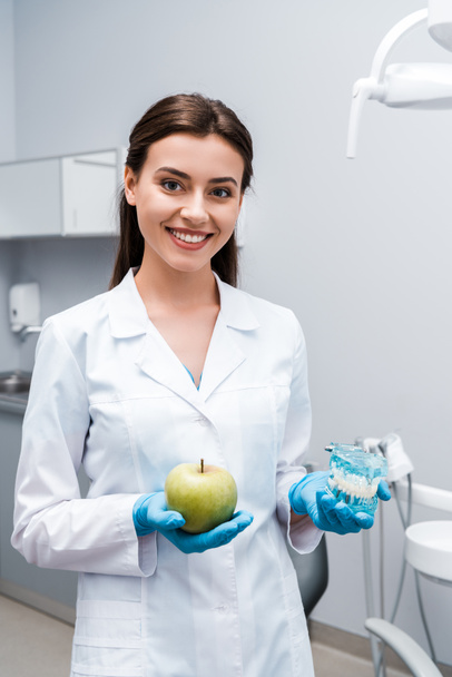 atractivo y feliz dentista sosteniendo los dientes modelo y sabrosa manzana en la clínica
  - Foto, Imagen