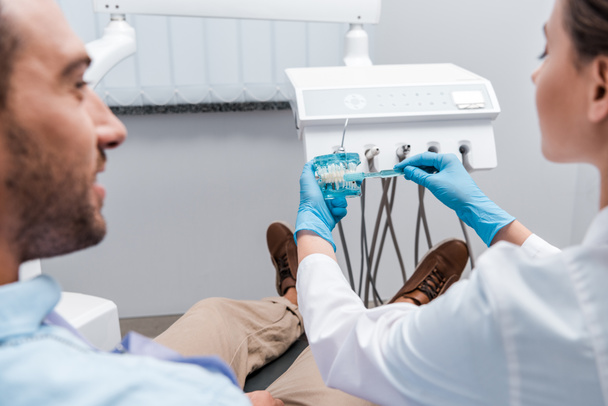 περικομμένη εικόνα του οδοντίατρου που κρατά οδοντόβουρτσα κοντά στα δόντια μοντέλο και ο άνθρωπος στην κλινική  - Φωτογραφία, εικόνα