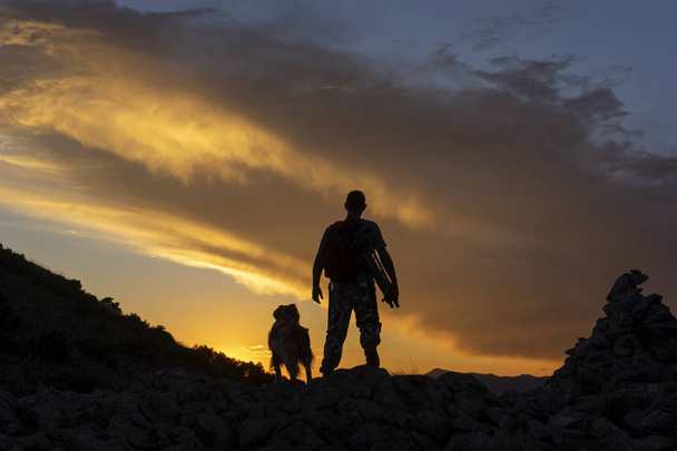 Ο πεζοπόρος που περπατάει με το σκύλο του στο ηλιοβασίλεμα στο βουνό - Φωτογραφία, εικόνα