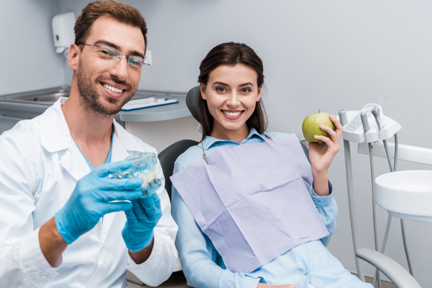 foyer sélectif du dentiste joyeux dans les lunettes tenant modèle de dents près de femme heureuse avec pomme verte
  - Photo, image