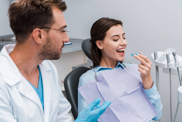 selective focus of cheerful woman looking at retainer near handsome dentist in glasses  - Foto, Bild