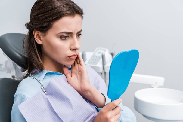 attractive and upset girl touching face while having toothache and looking at mirror in clinic  - Photo, Image