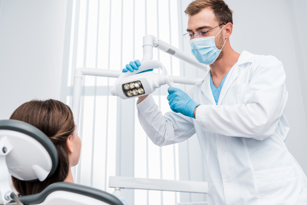 selektivní zaměření zubního lékaře v lékařské masce a v modrých latexových rukavicích dotýkání se zdravotnické lampy v blízkosti pacienta  - Fotografie, Obrázek