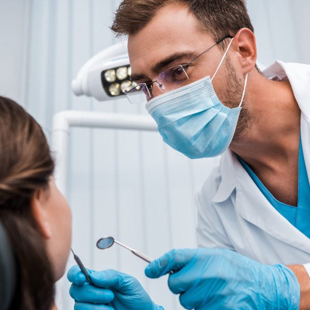 enfoque selectivo del dentista en gafas y máscara médica que sostiene instrumentos dentales cerca del paciente
  - Foto, Imagen