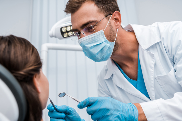 Selektiver Fokus des Zahnarztes in der medizinischen Maske, die zahnärztliche Instrumente in der Nähe des Patienten hält  - Foto, Bild