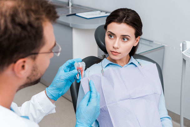 selective focus of young woman looking at dentist in latex gloves holding tooth model and dental instrument  - Φωτογραφία, εικόνα
