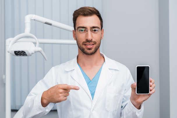 handsome bearded man in white coat pointing with finger at smartphone with blank screen  - Foto, Bild
