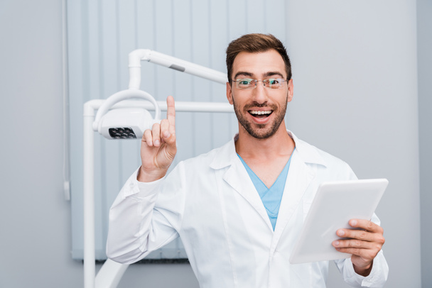 fröhlicher bärtiger Mann in weißem Mantel und Brille gestikuliert in der Hand eines digitalen Tablets  - Foto, Bild