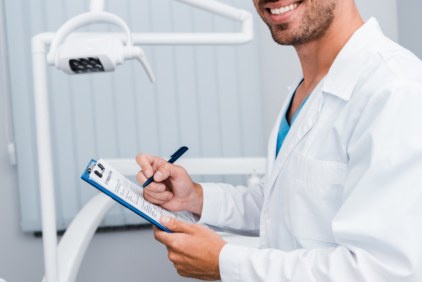 vista cortada do homem barbudo em branco casaco segurando caneta e área de transferência na clínica odontológica
  - Foto, Imagem