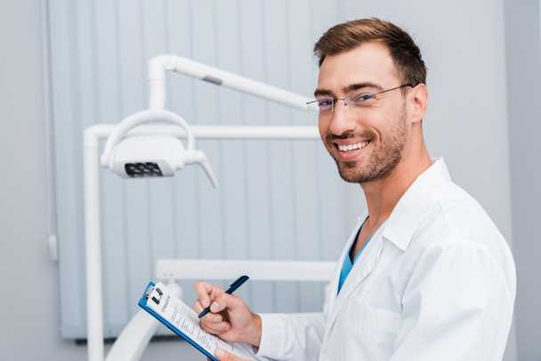 pohledný vousatý muž v bílém kabátě a brýlích s perem a schránkou na stomatologické klinice  - Fotografie, Obrázek