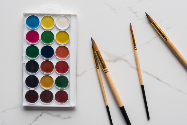 top view of watercolor paint palette and paintbrushes on marble white surface - Foto, afbeelding