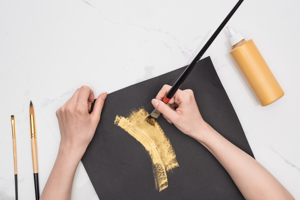 oříznutý pohled na kresbu výtvarníka se zlatou barvou v černém papíře na mramorovém povrchu - Fotografie, Obrázek