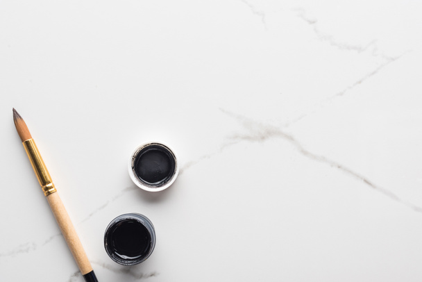 bovenaanzicht van zwarte gouache-verf en Paintbrush op marmer wit oppervlak met Kopieer ruimte - Foto, afbeelding