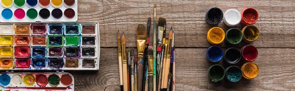 vista superior de coloridas paletas de pintura sobre superficie marrón madera con pinceles y gouache, plano panorámico
 - Foto, Imagen