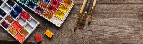vista dall'alto di tavolozze di colori ad acquerello su superficie marrone legno con pennelli, colpo panoramico
 - Foto, immagini